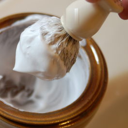 Shop detail: a shaving brush whips lather in a bowl, with a fingertip at the edge of the frame.