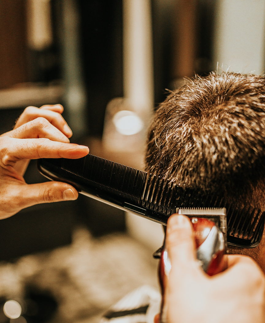 A barber holds a cutting comb flat against the back of a client's head and runs clippers along it. Photographed from behind; no face is in frame.