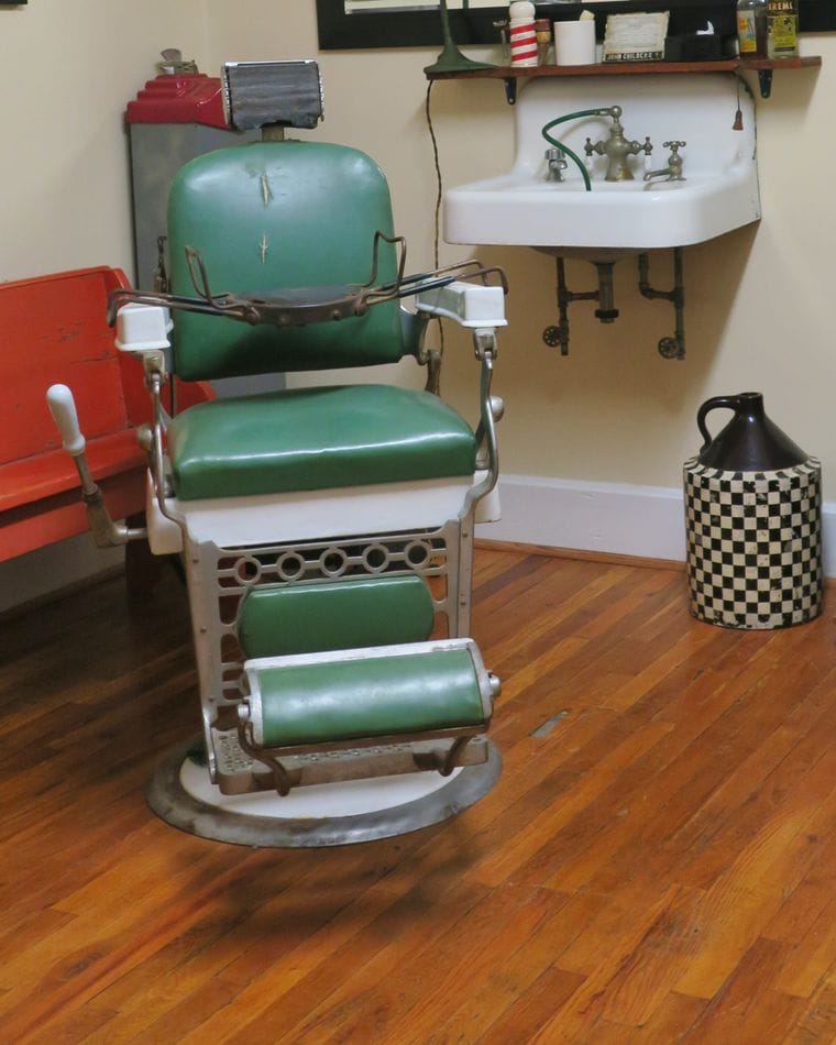 A green leather barber chair with a chrome footrest on a wooden floor, beside a white wall-mounted basin.