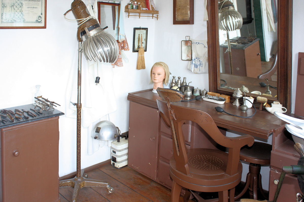 A hairdresser's room with a wooden counter, a mirror, a wig block and a chrome hood dryer on a stand.