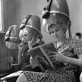 Salon detail: women reading under hood dryers in a black-and-white photograph.