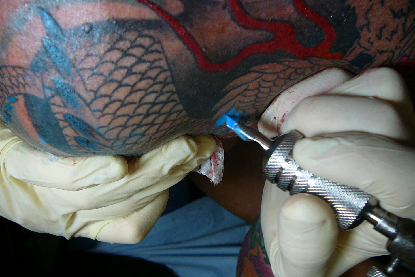 Gloved hands working colour into a large scaled backpiece with a coil machine.