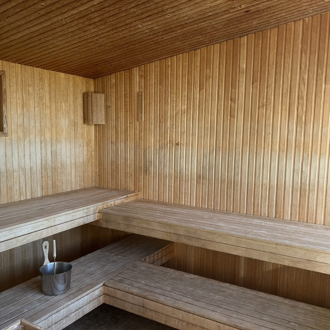 A cedar sauna with tiered benches along two walls, a metal pail on the lower bench and a small window at the end.