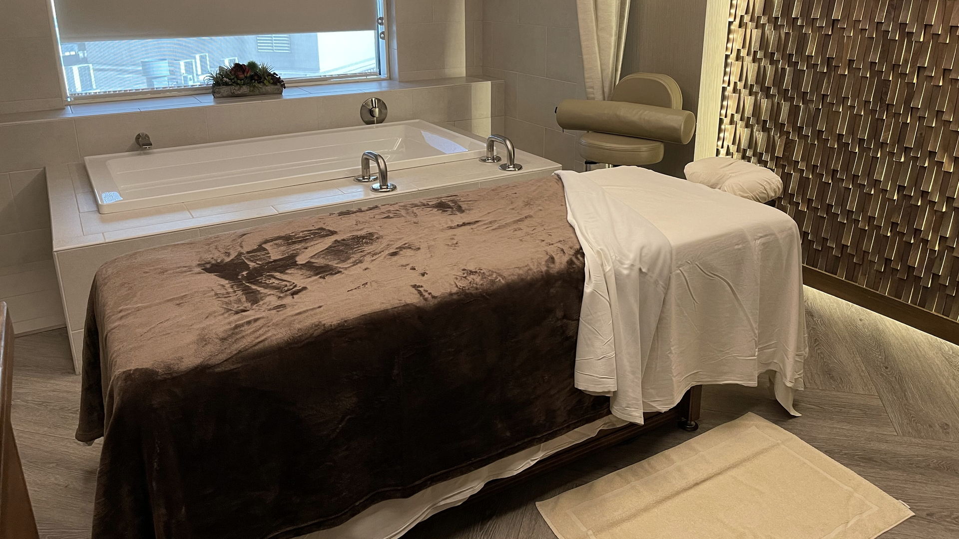 A treatment room with a massage table under a brown throw, a deep white bathtub, a window with a roller blind and a textured wood-tile wall.