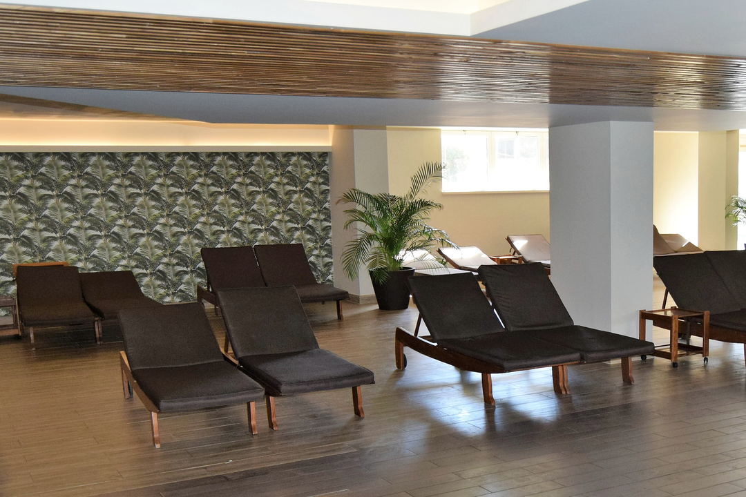 A lounge with rows of dark loungers, palm-leaf wallpaper, a potted palm, white columns and a slatted wood ceiling.