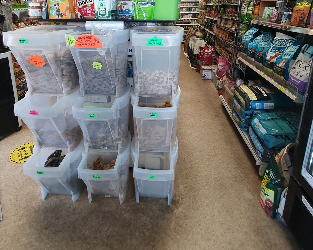 An aisle in a small pet supply shop, with stacked clear treat dispensers on one side and bagged dog food on the shelving opposite.