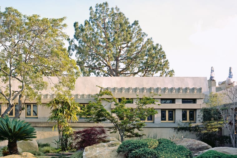 A long, low house with a moulded parapet, seen across a planted garden of boulders and trees.