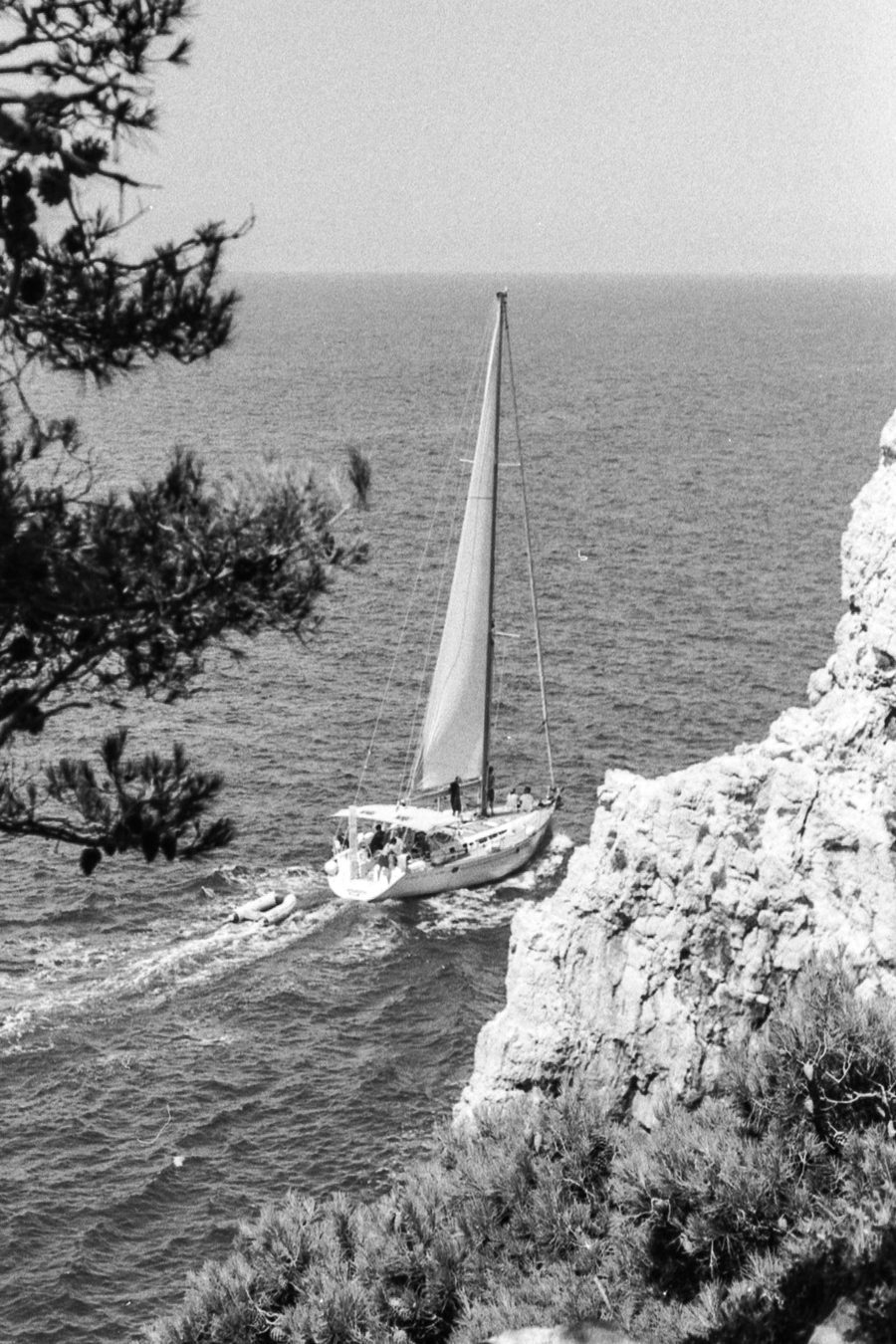 A sailing yacht under mainsail and jib, tender in tow, working close along a rocky limestone coast.