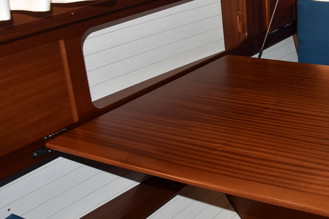A varnished mahogany saloon table beside white hull lining and a slatted cabin sole, inside a small sailing yacht.