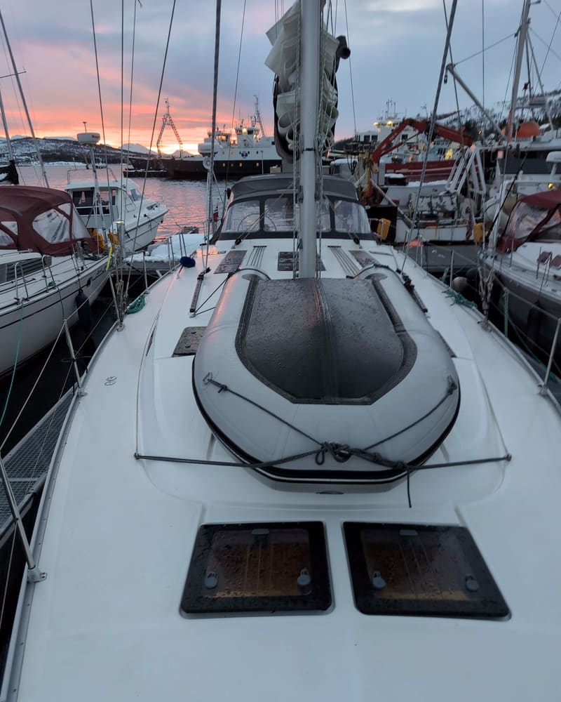 The foredeck of a sailing yacht moored in a marina at dusk, with a deck hatch and the mast rising into the evening sky.