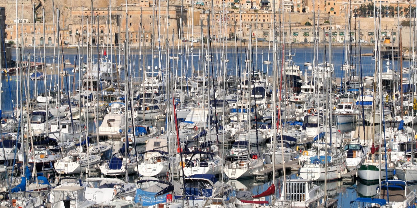 Rows of masted yachts moored along a marina quay beneath a stone harbour wall.