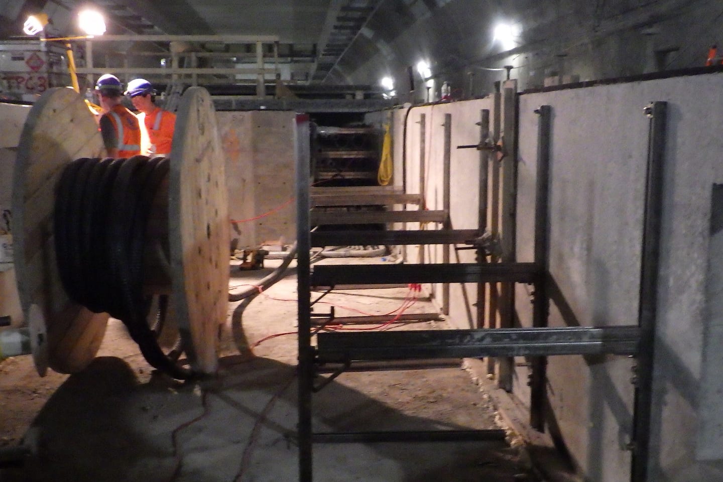 A cable drum and steel support frames set up along a concrete tunnel wall during a conduit installation