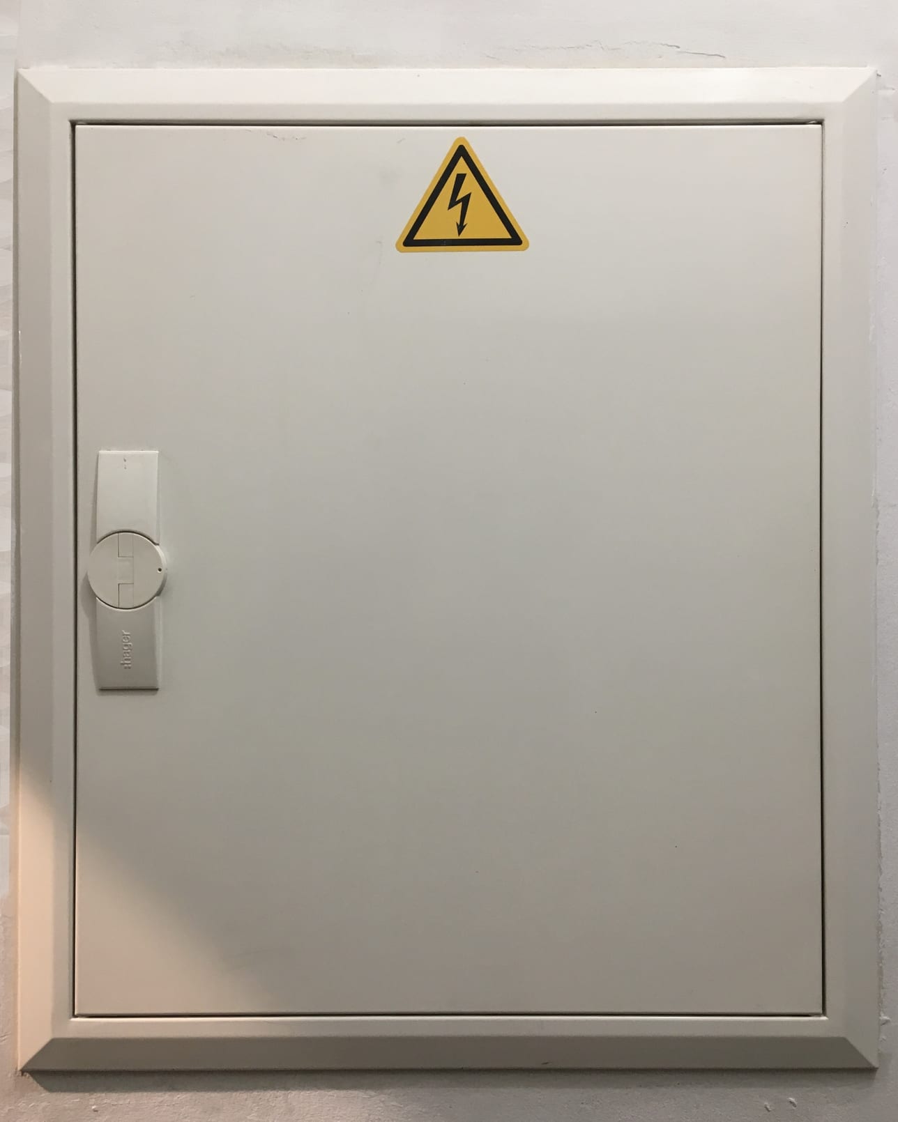 A closed metal distribution cabinet on a wall, its door carrying a yellow electrical hazard triangle.