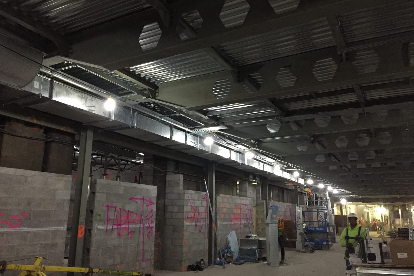 Overhead ductwork running the length of a corridor, hung on brackets beside lighting and pipe runs.
