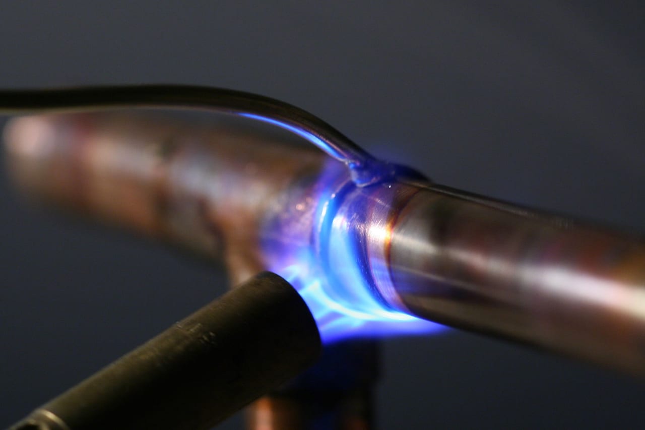 A propane torch flame held against a copper pipe joint, the solder seam bright at the shoulder of the fitting and the pipe run continuing past it