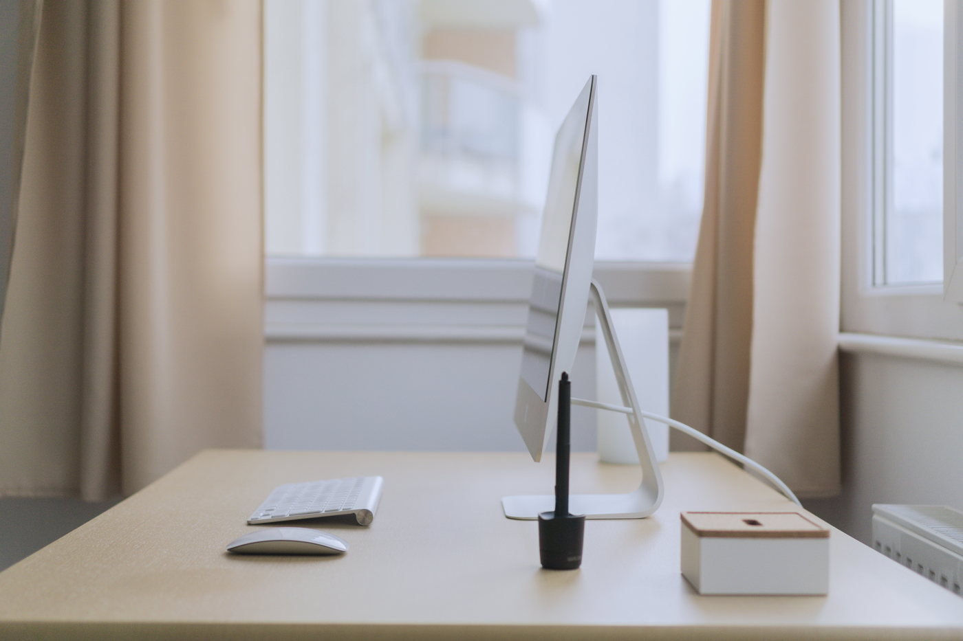 A pale wooden desk beside a curtained window, holding a computer monitor, a keyboard and a small wooden box.