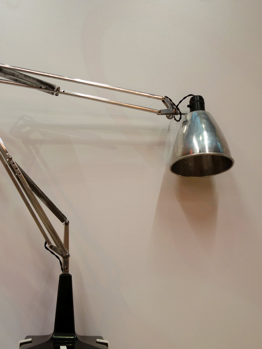 An articulated metal desk lamp with a spun shade, photographed against a plain pale wall.