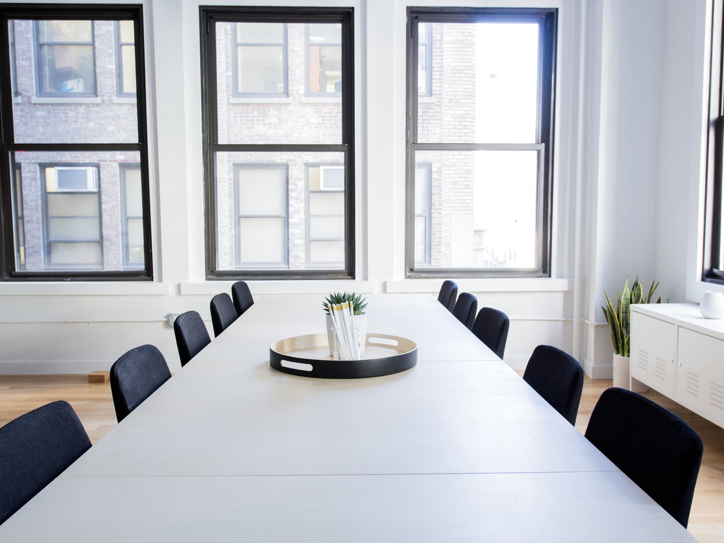 A long white meeting table with dark chairs in a bright room, lit by three tall windows facing a brick building.