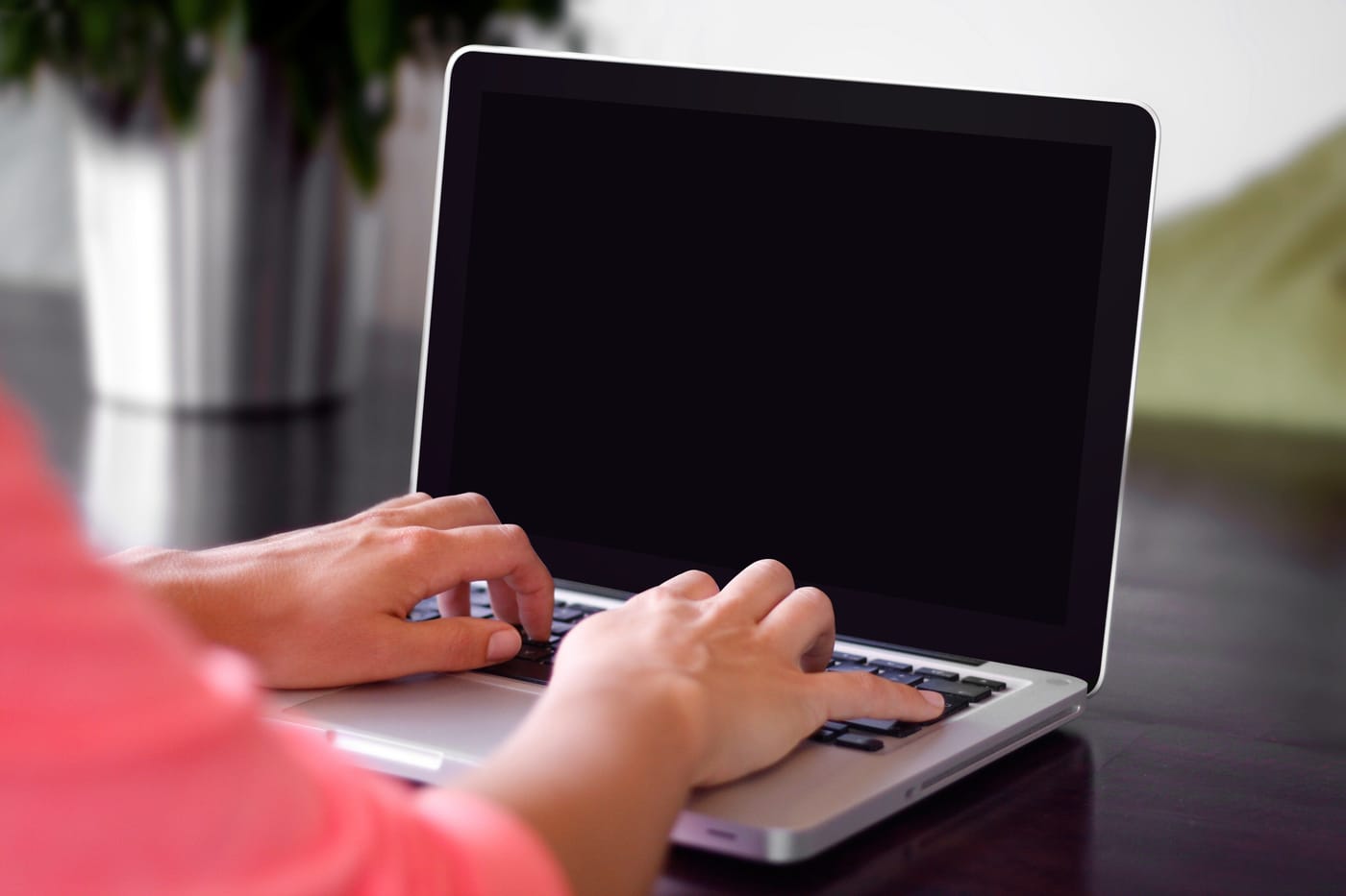 Hands in a coral sleeve typing on a laptop at a dark desk, beside a plant.