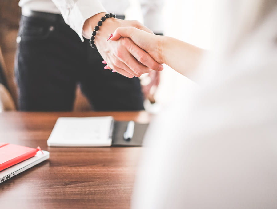 Two people shake hands across a meeting desk, an open notebook and a laptop on the table between them