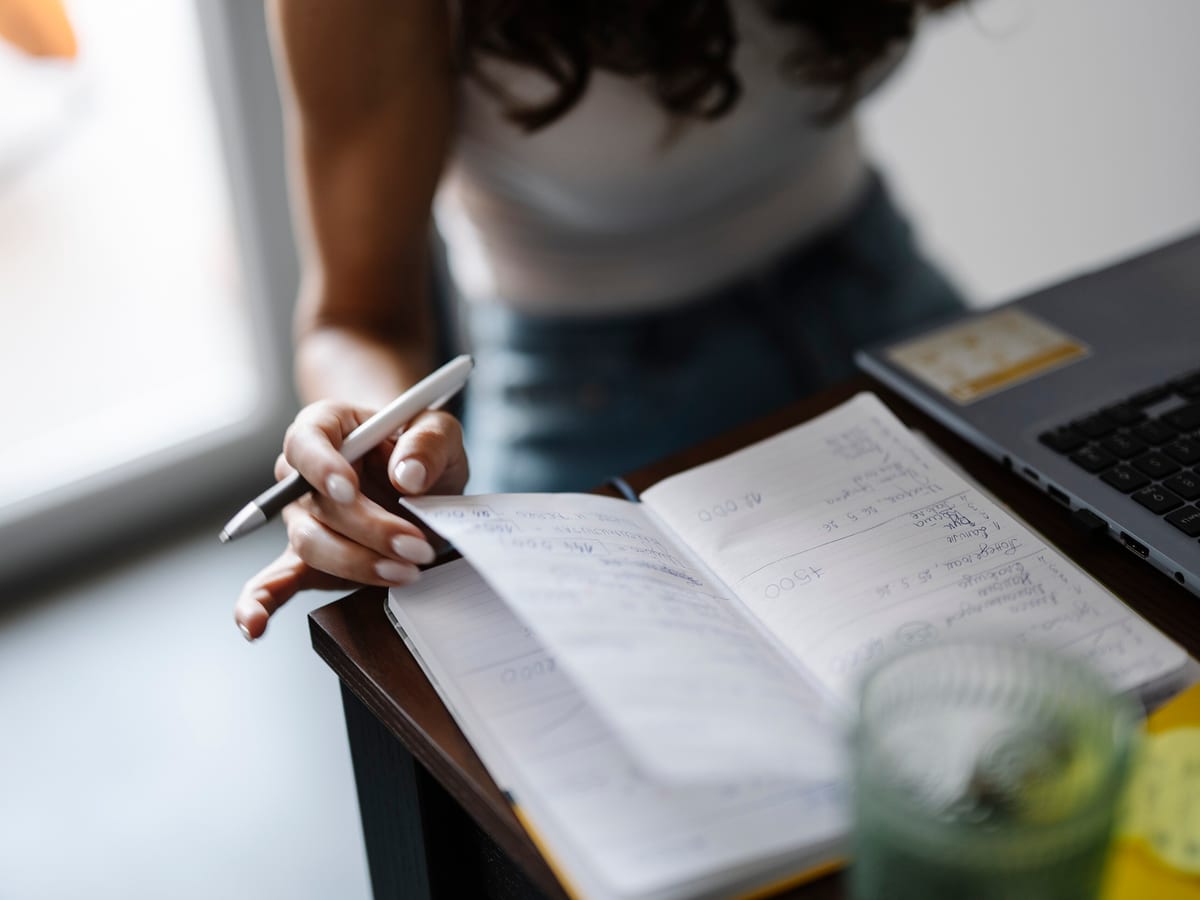 A hand writing in a notebook at a desk, photographed close enough that no face is shown.