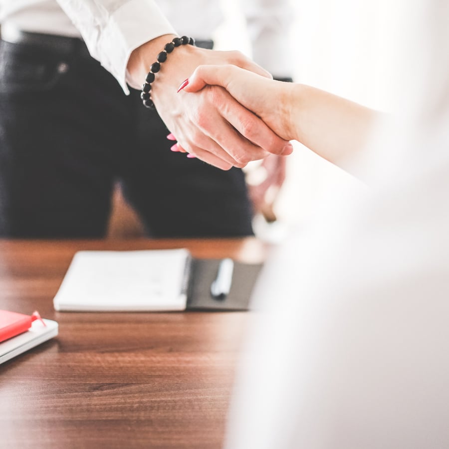 Two people shaking hands across a desk, with a notebook and pen on the table