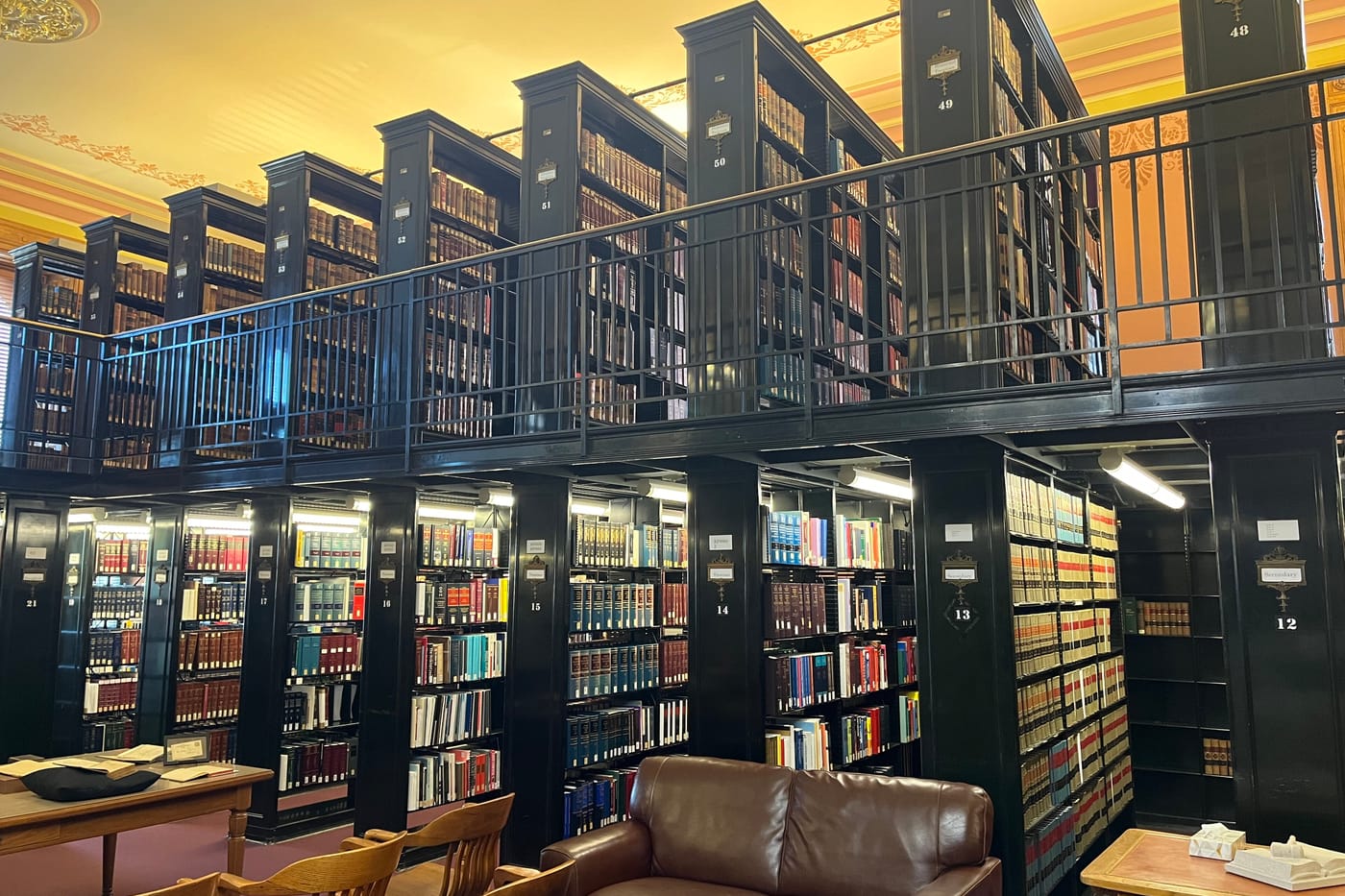 Two levels of dark steel bookstacks with a railed walkway above a leather reading chair.