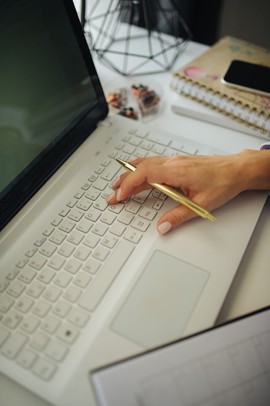 A hand holding a gold pen over a white laptop keyboard, with a notebook, a phone and a small plant on the desk beyond.