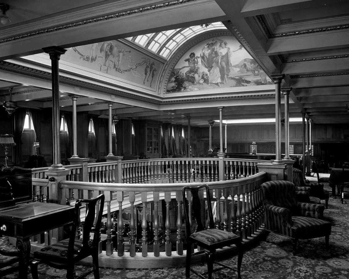 A long panelled saloon photographed in black and white, with armchairs and small tables set along both sides.