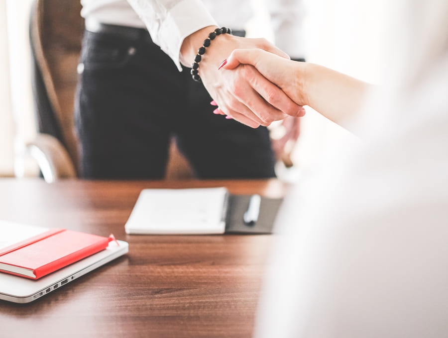 A handshake across a desk at the close of an advice meeting, notebook and laptop beside it