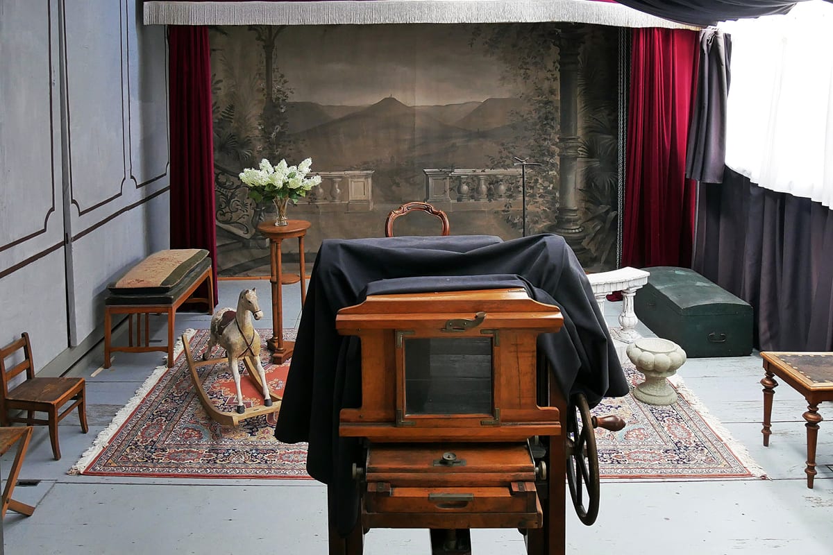 A reconstructed period photographic studio with a painted backdrop, a plate camera and a posing chair.