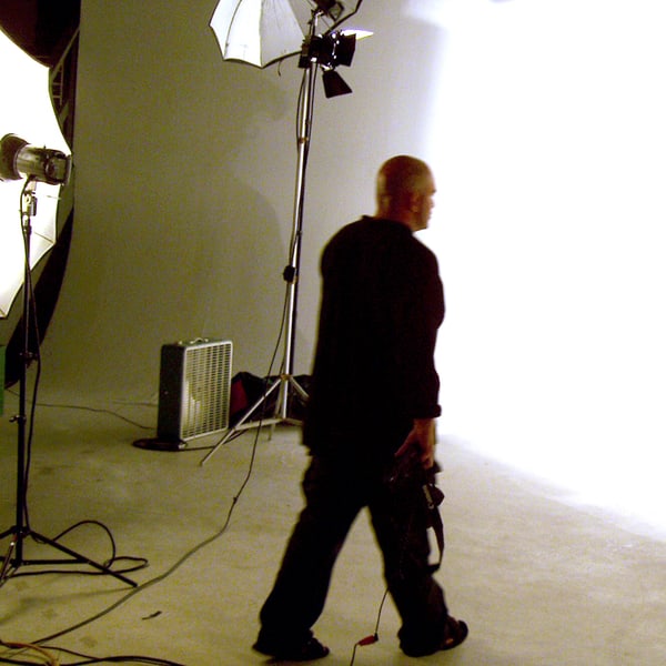 A photographer in silhouette crossing the lit white sweep, camera at his hip, umbrella and softbox burning either side
