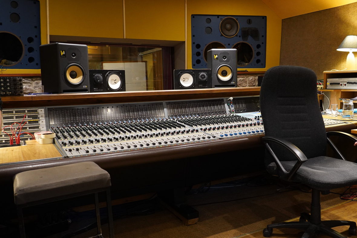 A control room with a long analogue console, monitor speakers above it and an empty chair at the desk.