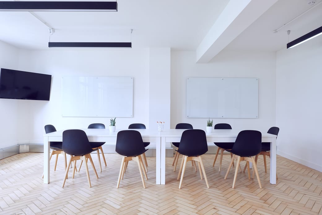 A long meeting table with dark chairs on a herringbone floor, lit by tall windows along one wall.