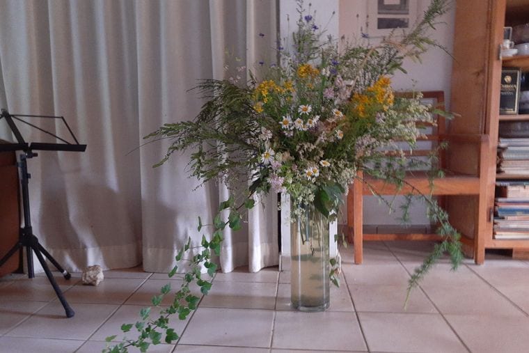 Wildflowers and trailing greenery arranged loosely in a tall glass vase on a tiled floor.