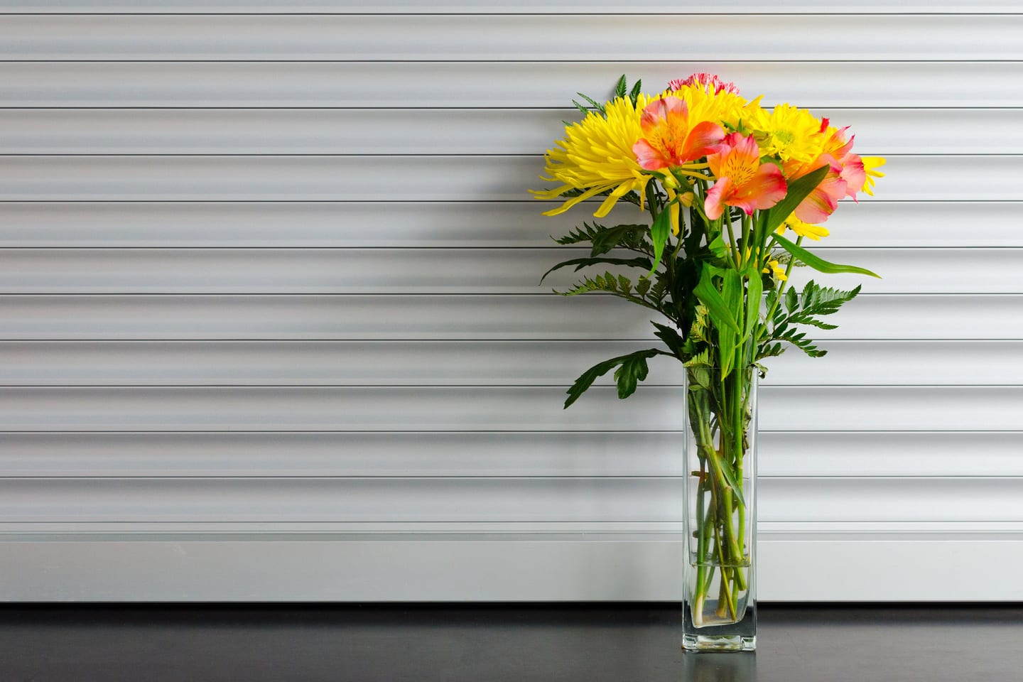 Orange and yellow gerberas in a slim glass vase against a white ribbed wall.