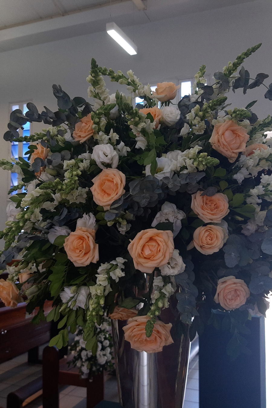 A tall arrangement of peach roses, white stocks and eucalyptus in a silver vase, standing beside wooden pews.