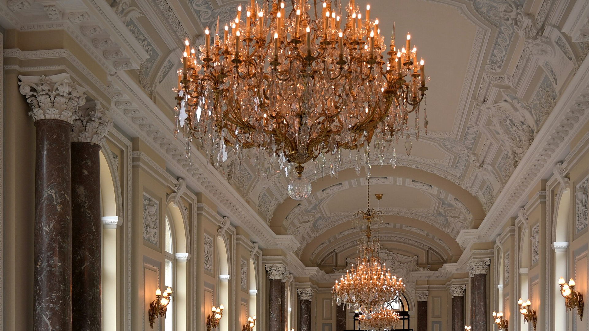 A long gallery with a moulded plaster ceiling, two gilt chandeliers hanging in line and dark marble columns down both sides.