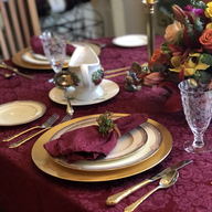 A burgundy cloth laid with gold chargers and gold cutlery, flowers behind it.