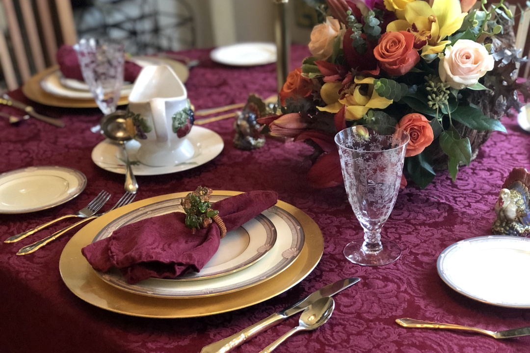 A place setting on a burgundy cloth: a gold charger, a folded napkin in a ring, gold cutlery and an etched glass goblet.
