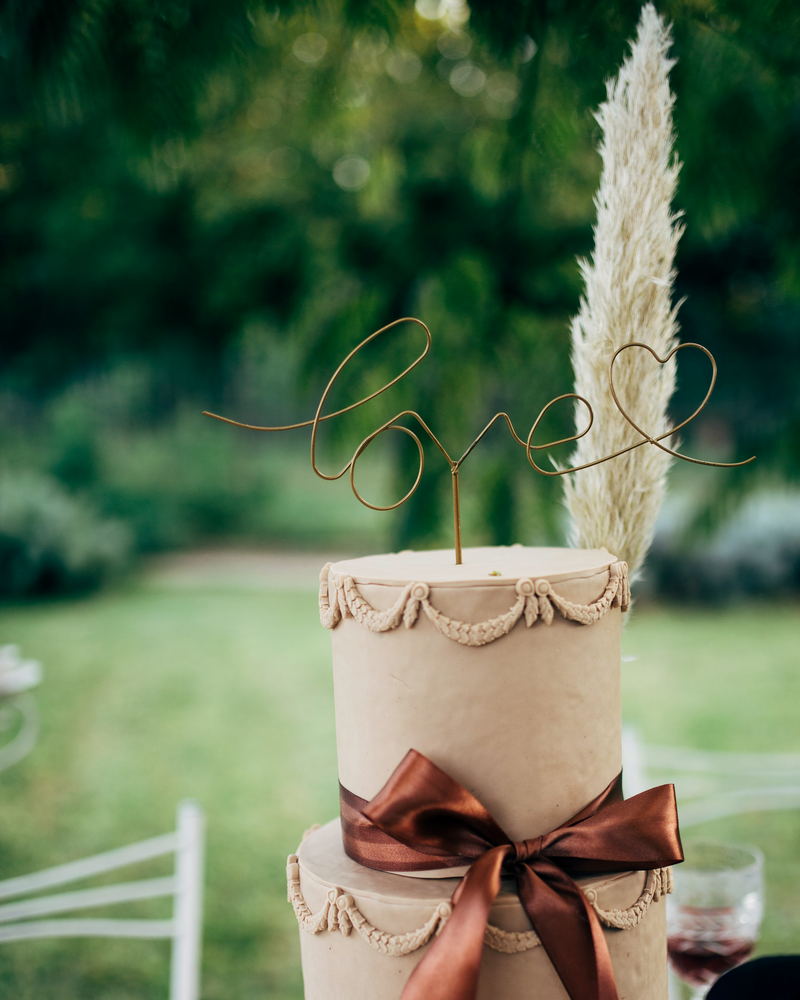 A two-tier cake standing on grass, with rope piping, a satin bow and a wire topper reading love.