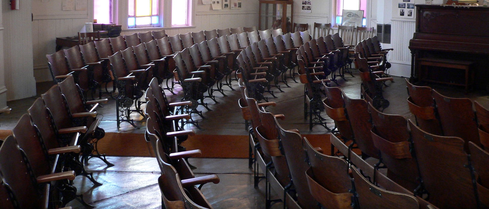 Curved rows of empty auditorium seats photographed from the back of the hall, with an upright piano standing at the side