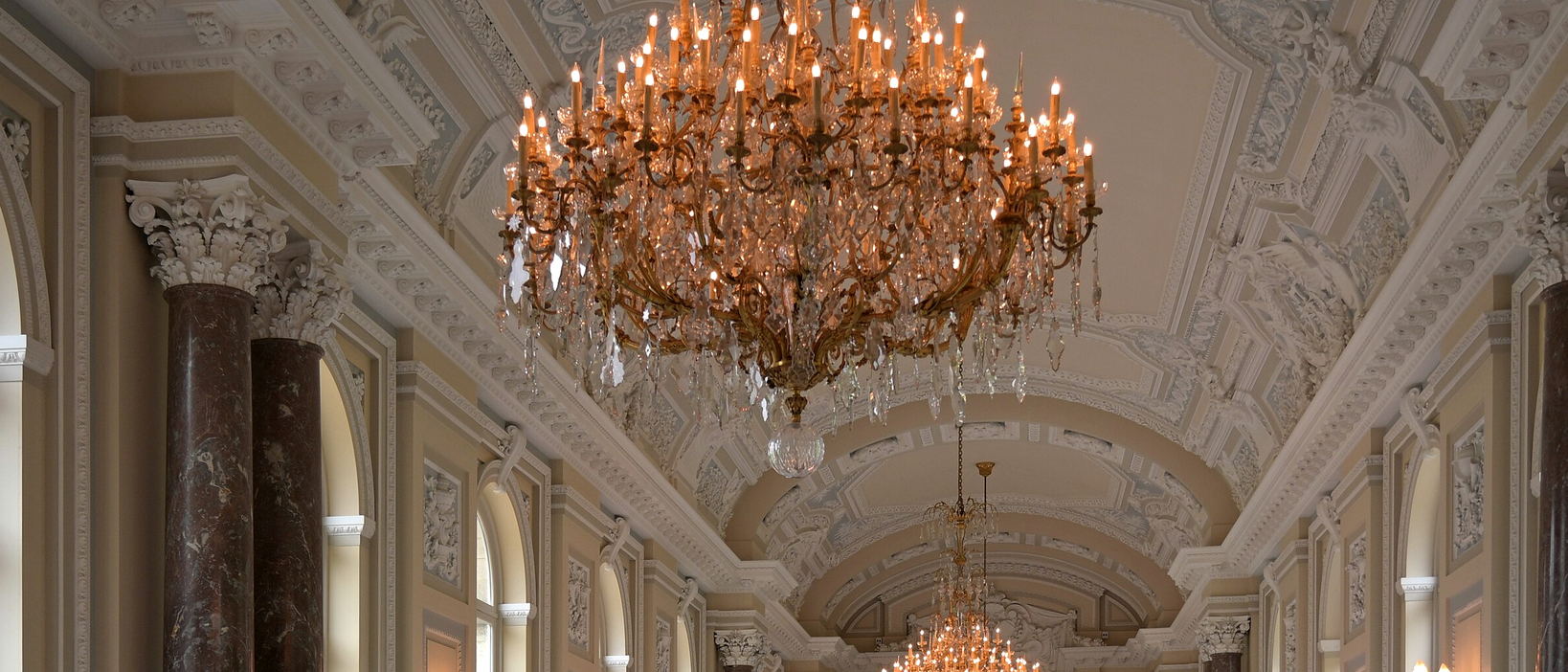 A gilt chandelier hanging close under a moulded plaster ceiling, with marble columns running away beneath it.