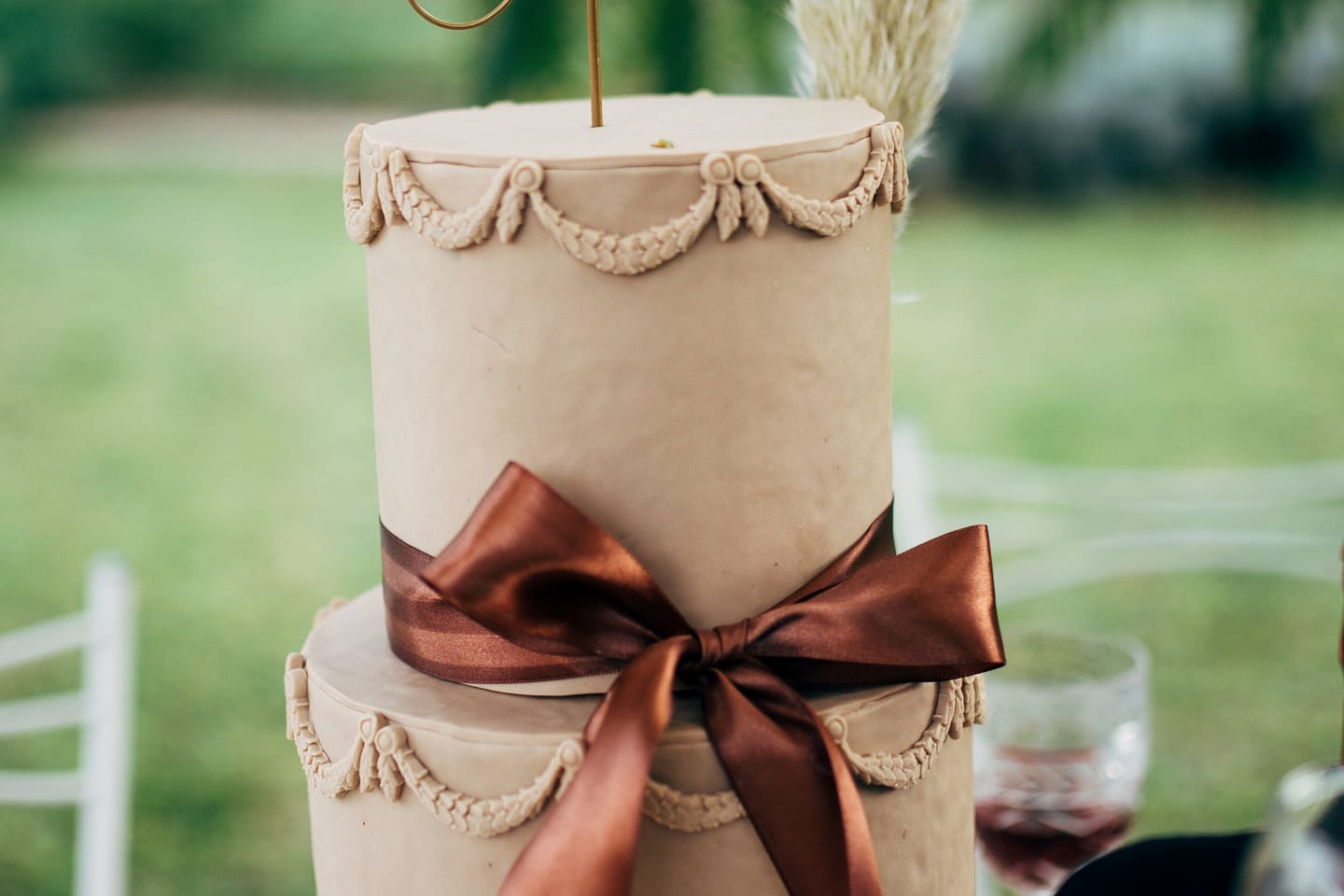 A two-tier pale wedding cake standing on grass, piped with rope detail and tied with a wide bronze satin bow.