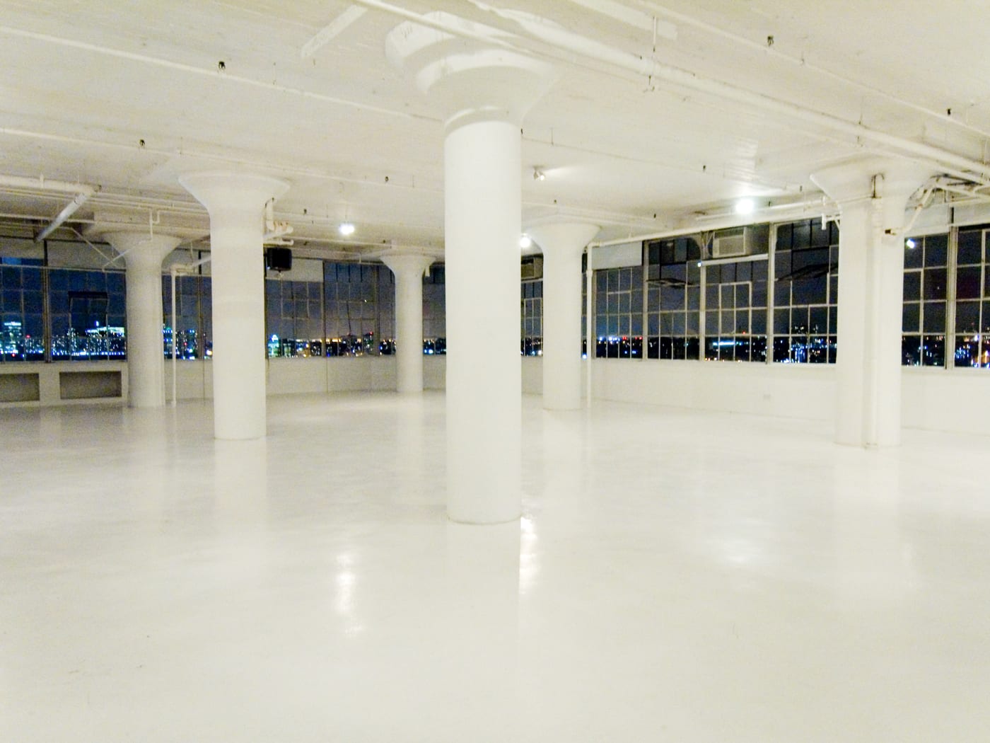 An empty white loft floor with round columns and tall windows looking out on a city skyline at night.