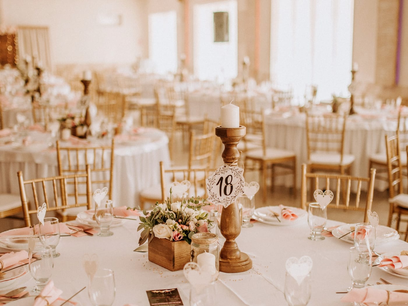 Round reception tables under white cloths with blush napkins, gold chairs and a table number on a turned wooden stand.