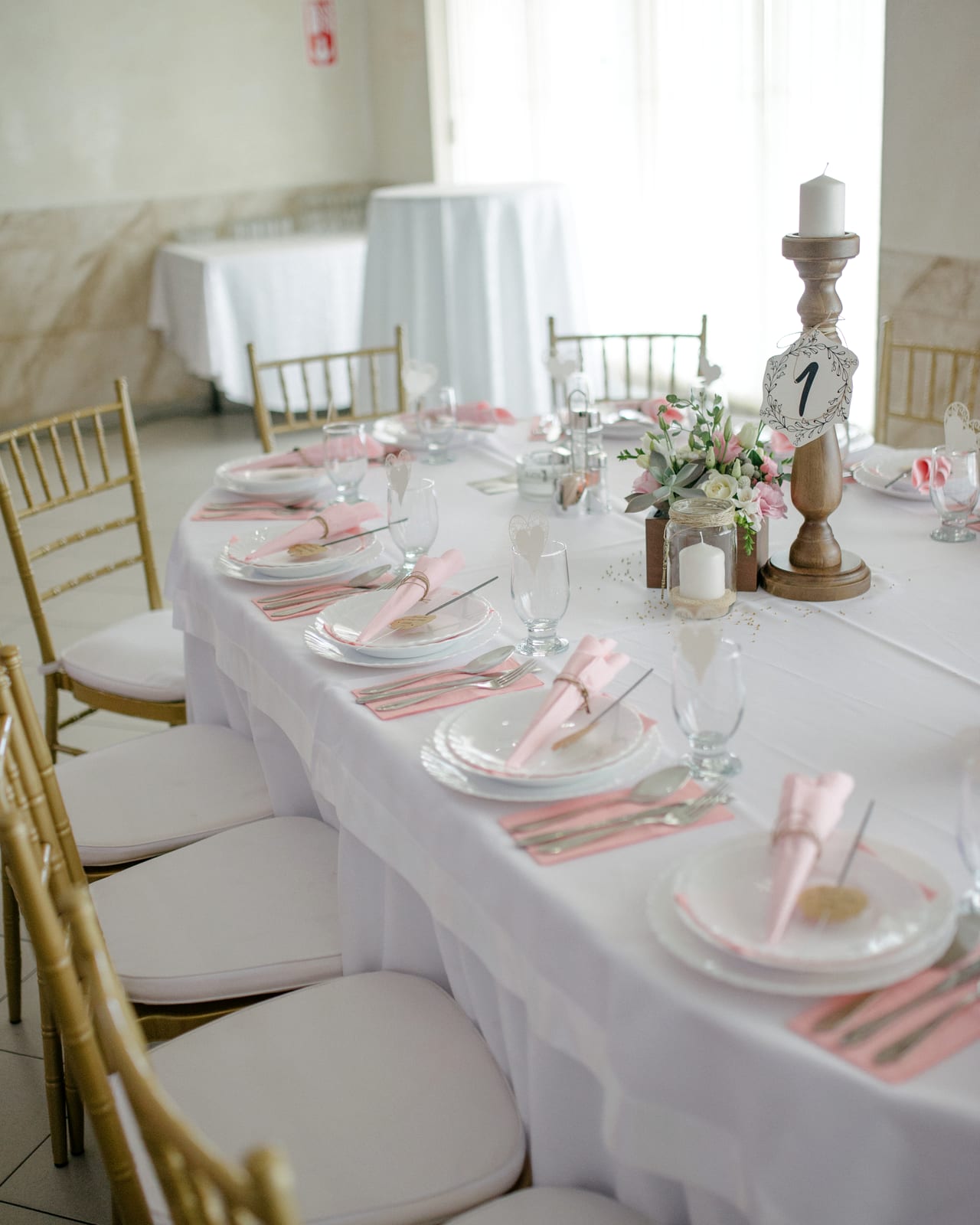A long table under a white cloth with blush napkins, gold chairs and table number one on a turned wooden stand.
