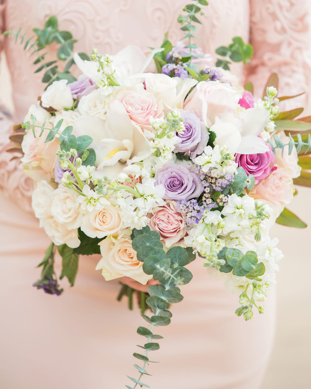 A bouquet of cream, peach and lilac roses with white stocks and trailing eucalyptus, held against a pale pink dress.