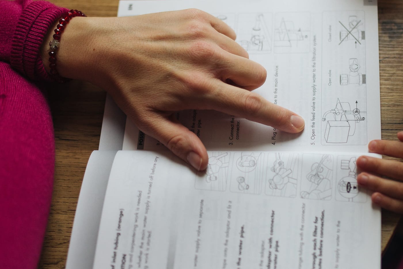 An adult hand pointing to a numbered step on a printed page of diagrams while a younger hand holds the edge of the booklet.