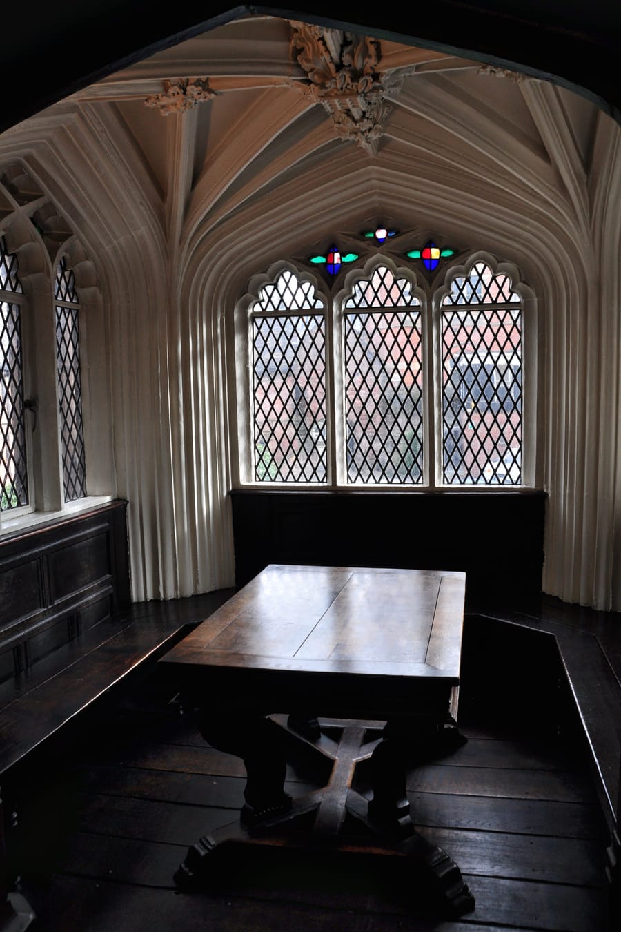 A heavy dark wooden table in a panelled reading alcove below a leaded bay window in an old library.